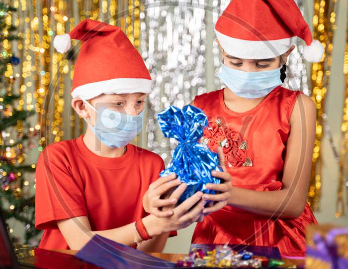 Kids In Medical Mask In Front Of Laptop Opening Gift At Home With Decorated Background During Christmas Eve - Concept Of Distant Xmas Celebration Due To Coronavirus Or Covid-19 Pandemic.