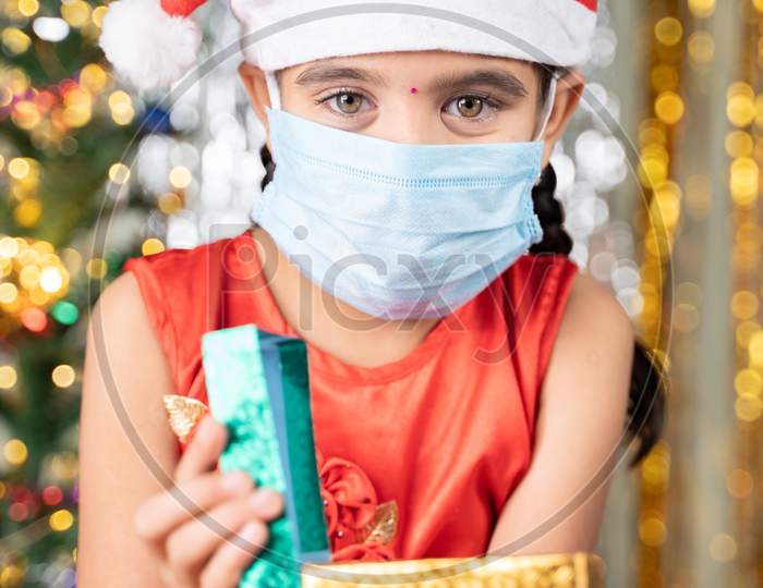 Girl Kid In Medical Mask With Christmas Hat And Decorated Background Opening Gift Box - Concept Of Gift Sharing, Distant Xmas Celebration Due To Ccoronavirus Or Covid-19 Pandemic.