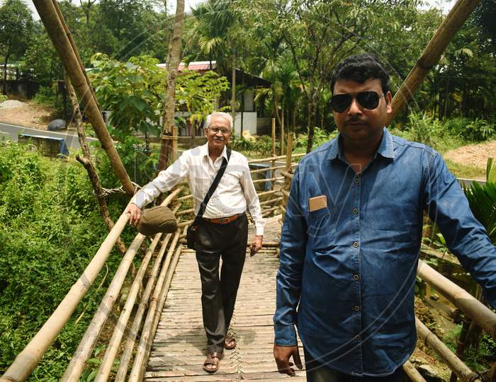Two Men Are Standing On A Bamboo Bridge In Mawlynnong, East Khasi Hills, Meghalaya, India. The Young And Old Persons, Wearing Blue And White Shirts, Are Looking Towards The Camera With Pose And Style.