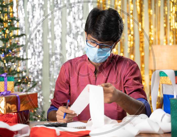 Young Man In Medical Mask Busy In Calculating Holiday Expenses Bills With Christmas Decorated Background And Gifts Infront Of Table.