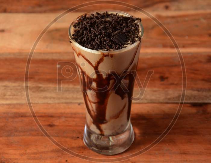 Cold Coffee In A Tall Glass With Chocolate Crushes Poured On A Old Rustic Wooden Table.