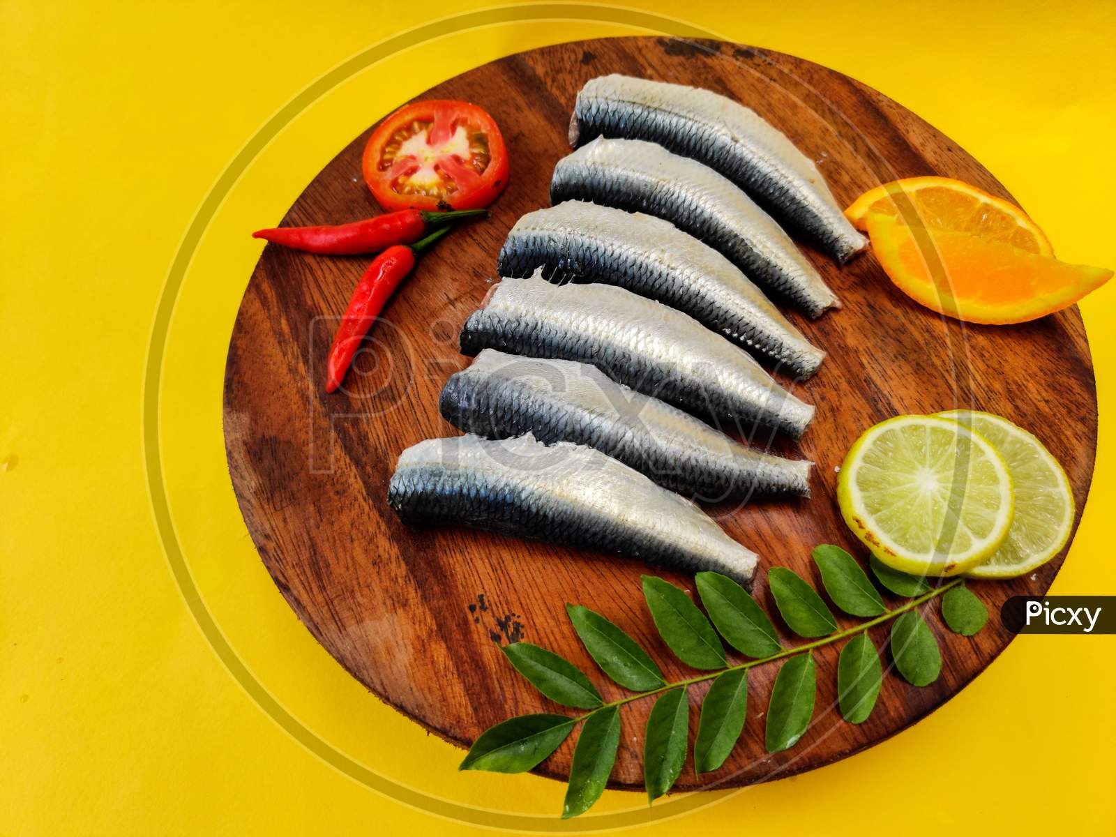 Cleaned And Ready To Cook Fresh Indian Oil Sardine (Sardinella Longipces) On A Wooden Pad With Curry Leaves,Tomato Slice And Red Chilli.Isolated On Yellow Background.
