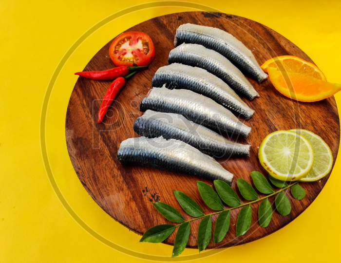 Cleaned And Ready To Cook Fresh Indian Oil Sardine (Sardinella Longipces) On A Wooden Pad With Curry Leaves,Tomato Slice And Red Chilli.Isolated On Yellow Background.