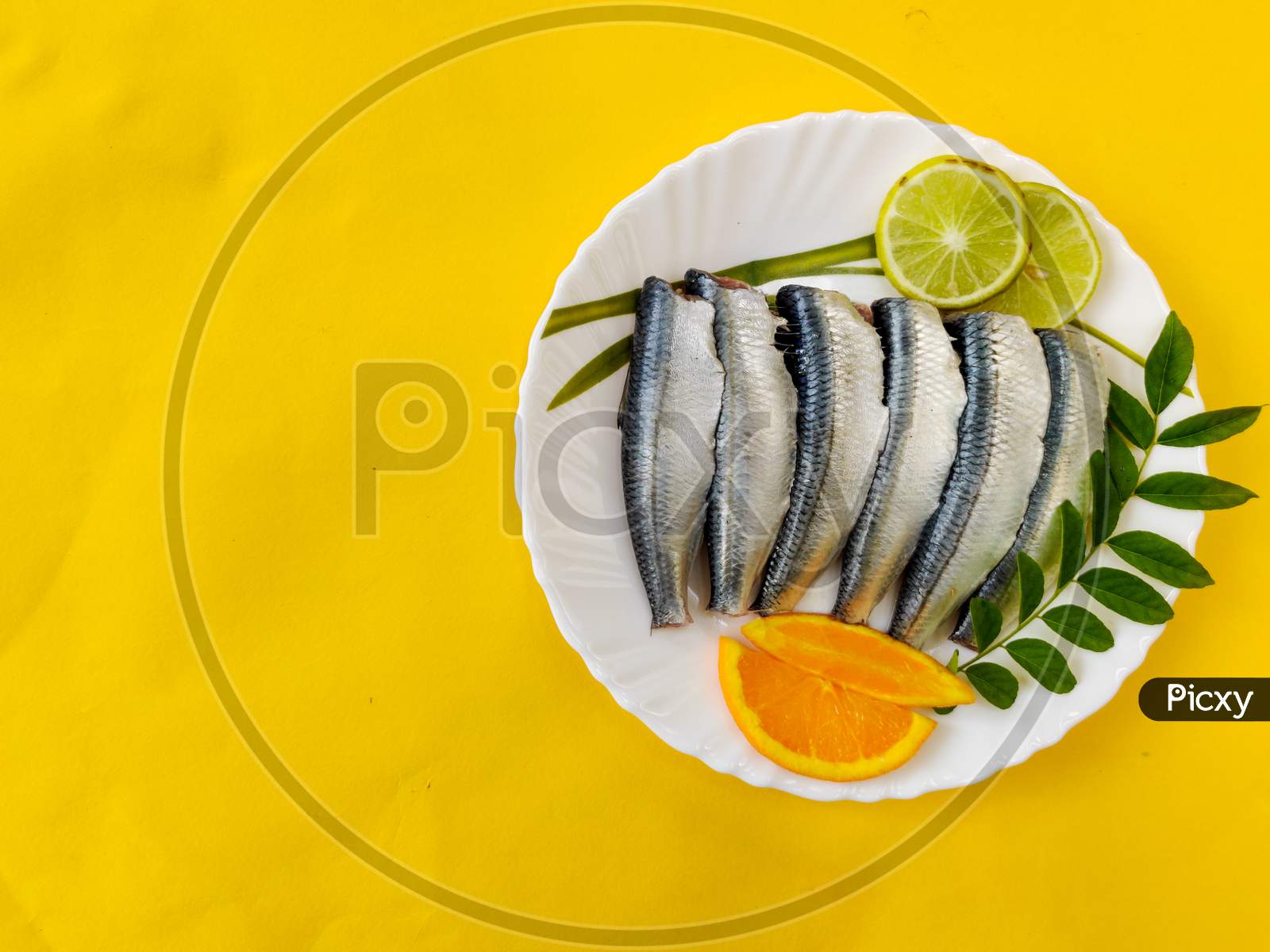 Cleaned And Ready To Cook Fresh Indian Sardine Decorated With Curry Leaves,Lemon Slice And Tomato Slice .Isolated On Yellow Background.