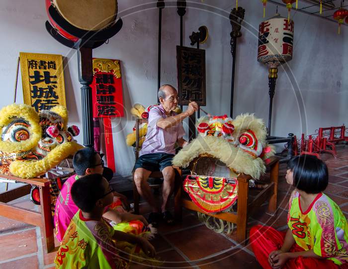 An Old Man Clean The Chinese Lion Dance Head With Brush With Teenagers Sit At The Floor
