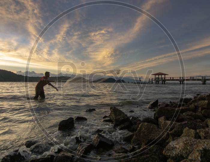 Fisherman Cast Net Near Rock Coastal In Sunset Hour