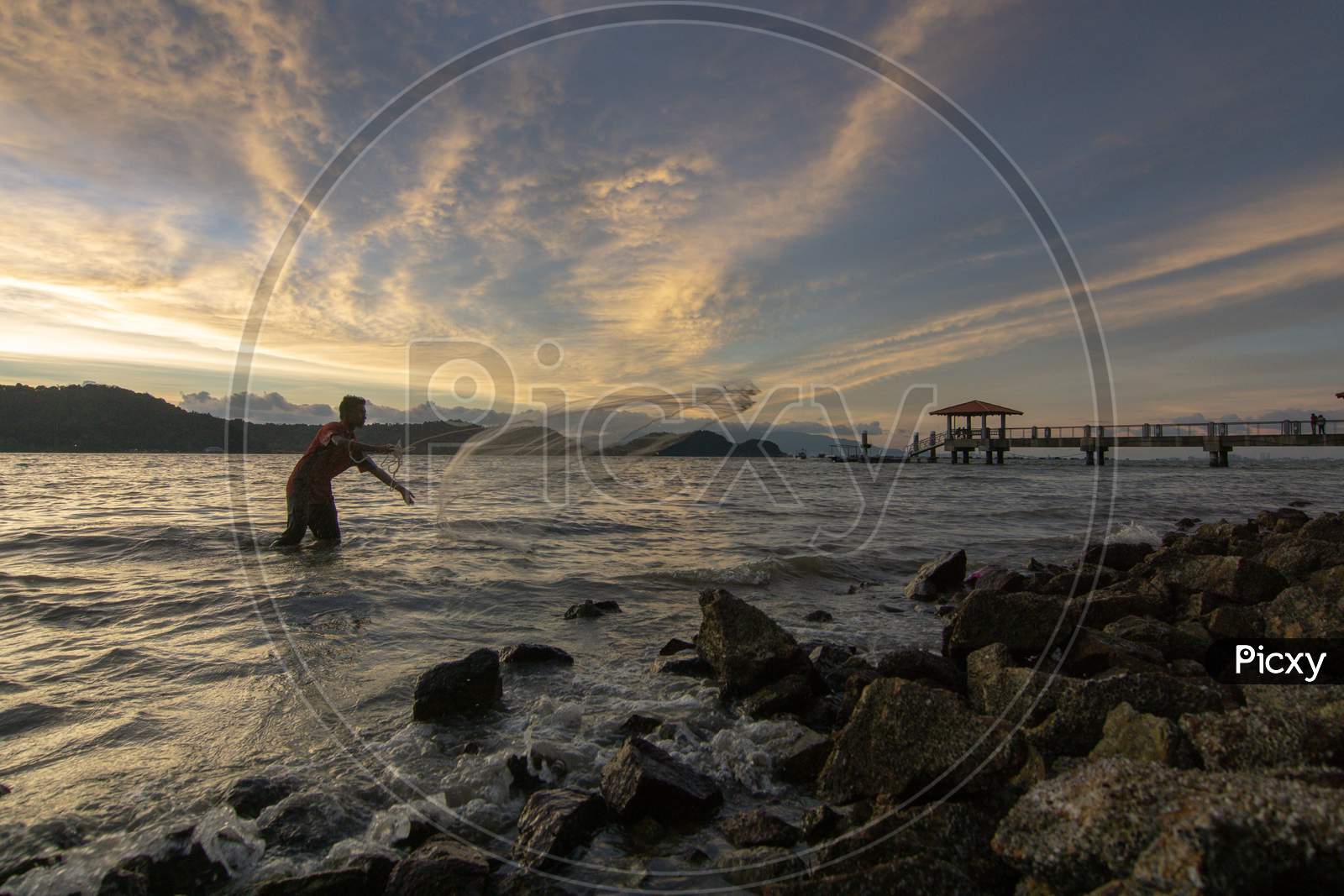 Fisherman Cast Net Near Rock Coastal In Sunset Hour