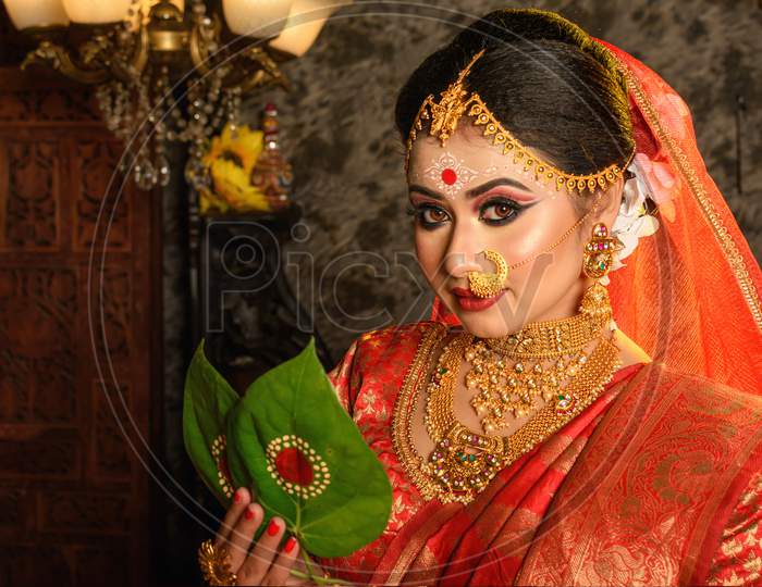 Portrait of very beautiful Indian bride holding betel leaf, Bengali bride in traditional wedding saree with makeup and heavy jewellery in studio lighting indoor