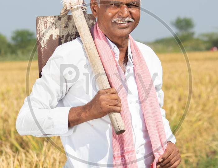Happy Smiling Indian Farmer With Hand Hoe Or Garden Spade Standing In The Middle Of Agriculture Farmland During Harvesting Season.