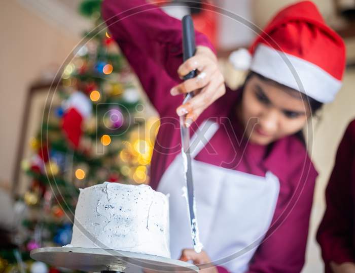 Young Indian Girl Icing Preparing Christmas Cake With Buttercream At Home, Low Angle Shot, Wear Santa Hat, Holiday And Party Season, Celebration Christmas During Covid pandemic. Cake and pastry Shop.