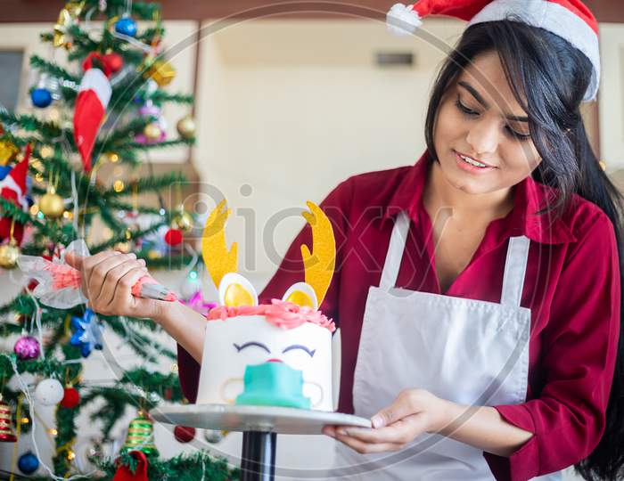 Young indian girl wearing apron preparing christmas deer design cake at home, celebration during covid pandemic, festive holiday season