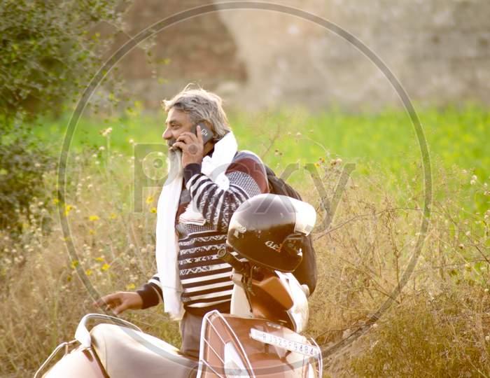 Old Bearded Farmer Man Talking Animatedly On A Mobile Phone Near His Farm Feild While A Two Wheeler With Helmet Stands Near Him Showing The Progress And Connectivity Of Rural India