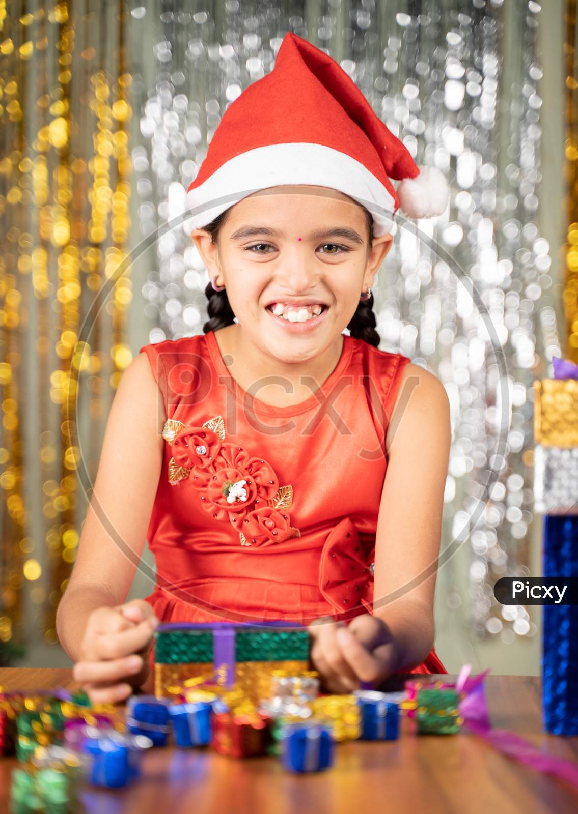 Happy Smiling Young Girl Kid With Santa Hat On Christmas Decorated Background Opens Gift Box During Holiday Celebration While Looking Camera.