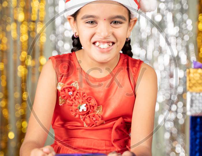 Happy Smiling Young Girl Kid With Santa Hat On Christmas Decorated Background Opens Gift Box During Holiday Celebration While Looking Camera.