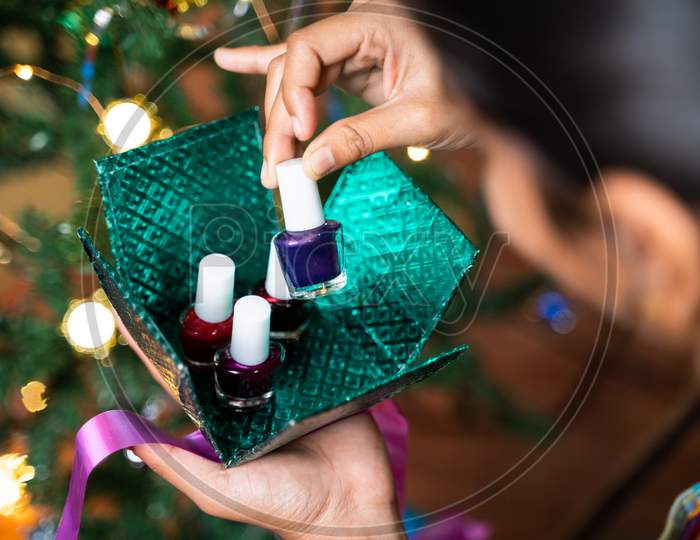 Concept Of Self Gifting During Holiday Shopping Or Celebration - Shoulder Shot Of Girl Infront Of Decorated Christmas Tree Opening Gift Box And Got Nail Polish Or Enamel As Gift.
