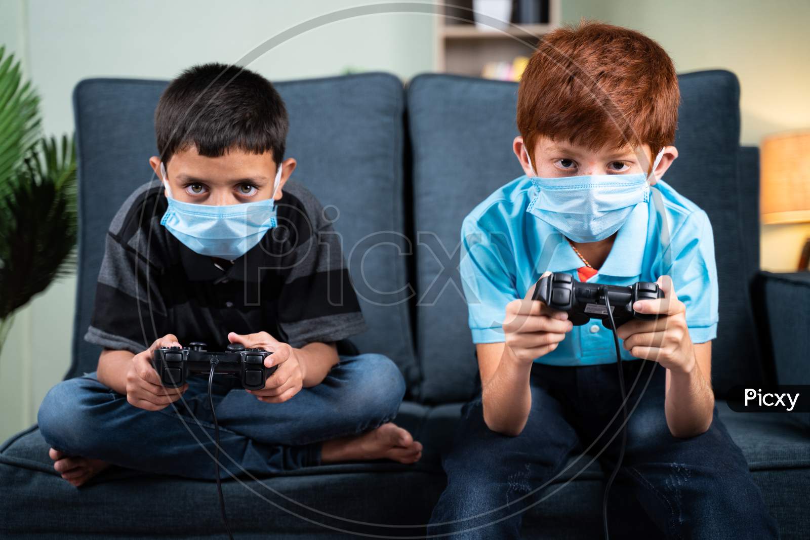 Two Kids With Medical Face Mask Seriously Busy Playing Video Game Using Joystick At Home - Concept Of Kids On Game During Coronavirus Covid-19 Lockdown.