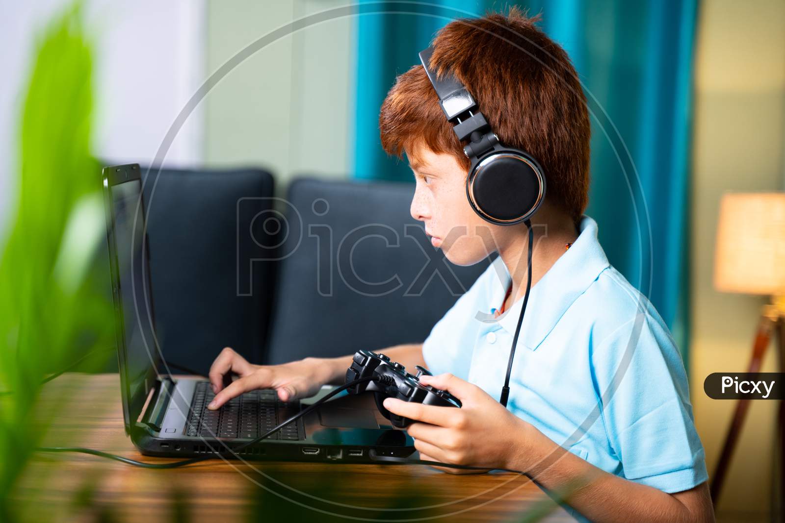 Kid Busy Searching And Playing Online Videogame On Laptop By Using Gamepad Or Joystick At Home By Wearing Headphone - Concept Of Kid Using Technology And Modern Lifestyle.
