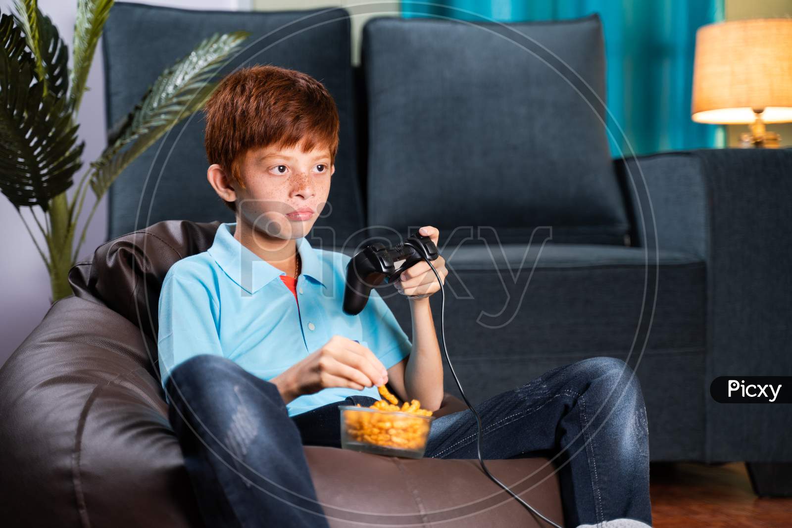 Kid Sitting On Bean Bag Seriously Playing Video Game While Eating Chips At Home During Leisure Time - Concept Of Kids Game Concentration And Lifestyle.