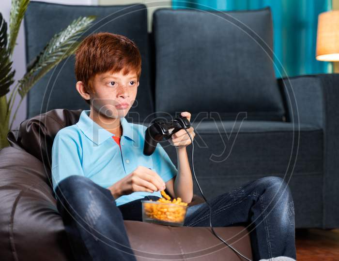 Kid Sitting On Bean Bag Seriously Playing Video Game While Eating Chips At Home During Leisure Time - Concept Of Kids Game Concentration And Lifestyle.
