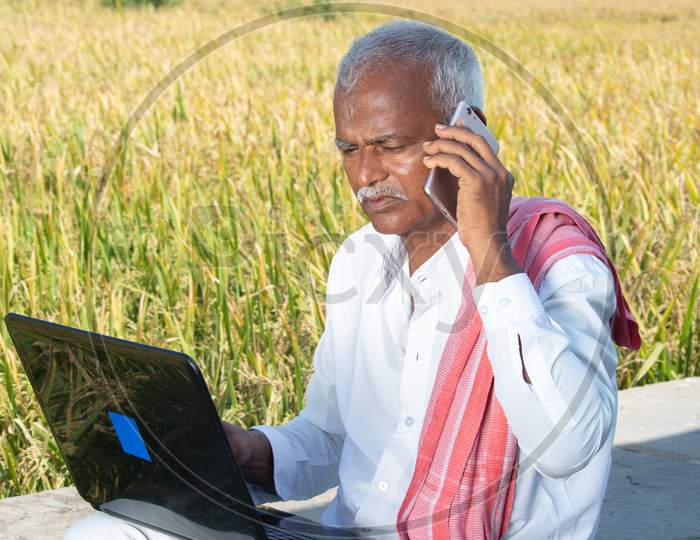 Indian Farmer Talking On Mobile Phone While Busy Looking Into Laptop Near The Agriculture Farmland - Concept Of Farmer Using Technology, Internet In Rural India.