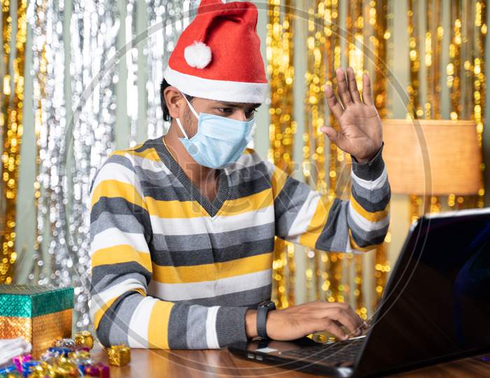 Young Man In Medical Mask On Video Call In Laptop During Christmas Or New Year Celebration Eve With Decorated Background And Gifts In Front - Concept Of Work From Home During Holydays.