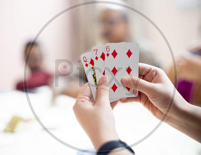 Woman Holding Cards With Out Of Focus Family Members In The Distance Showing A Family Playing Cards On Festivals Like Diwali, Dussera, New Year And More