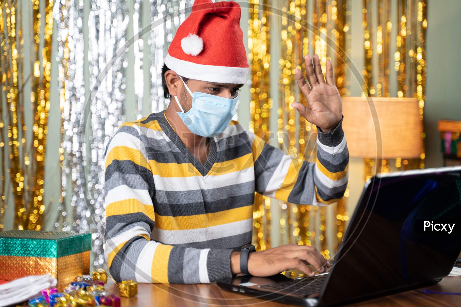 Young Man In Medical Mask On Video Call In Laptop During Christmas Or New Year Celebration Eve With Decorated Background And Gifts In Front - Concept Of Work From Home During Holydays.