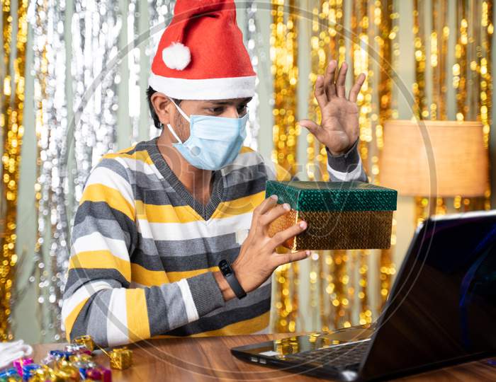 Young Man In Face Mask Showing Gift Box During Online Video Chat On Laptop During New Year Or Christmas Holiday Celebration.
