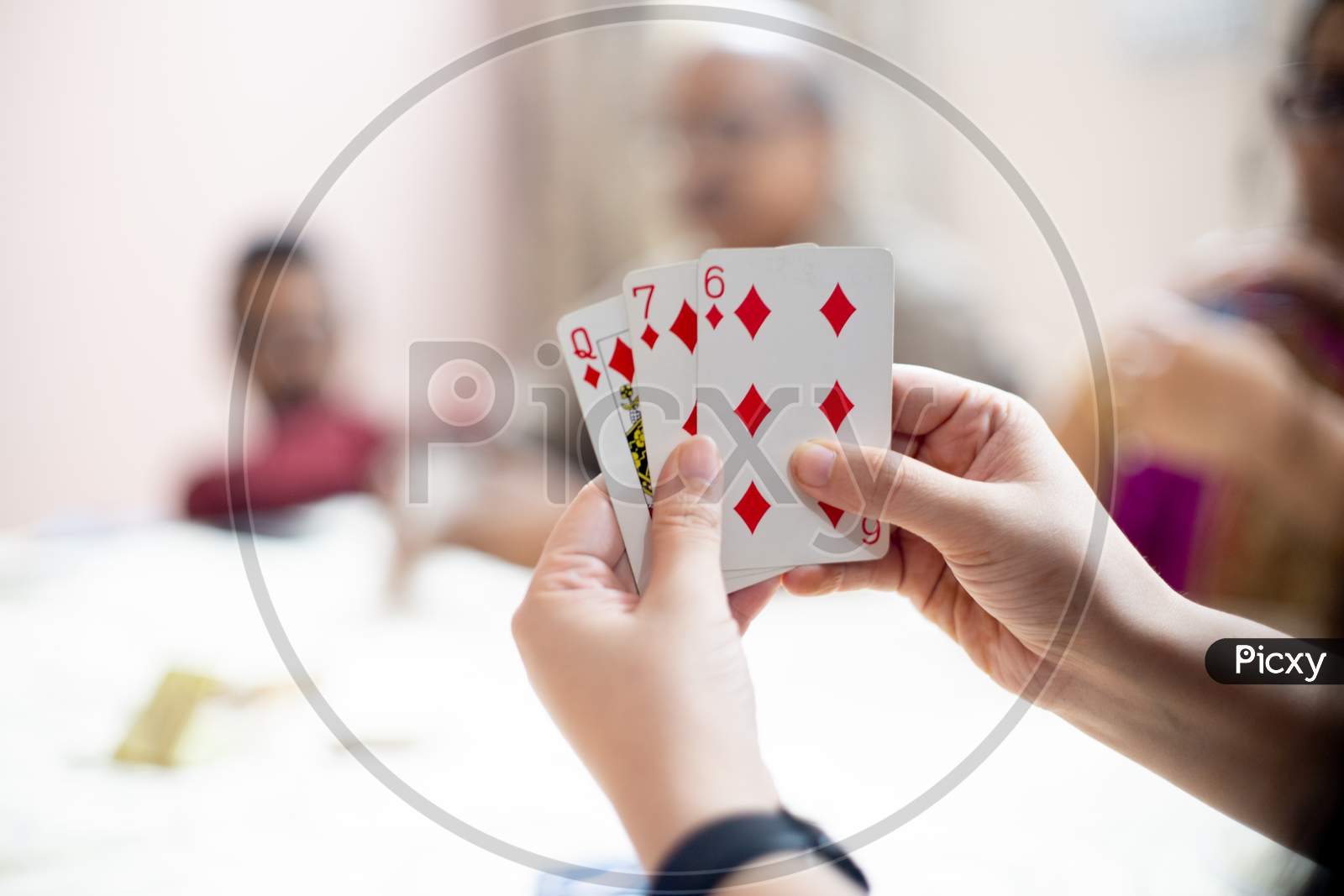 Woman Holding Cards With Out Of Focus Family Members In The Distance Showing A Family Playing Cards On Festivals Like Diwali, Dussera, New Year And More
