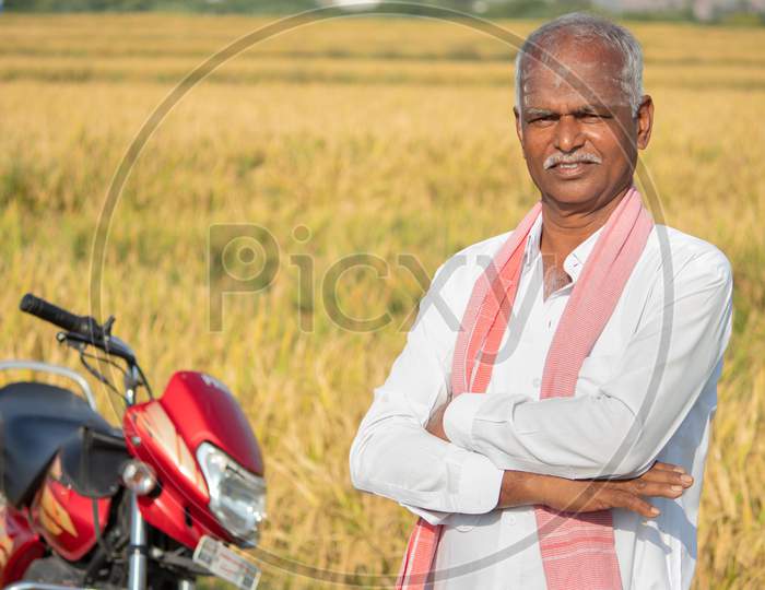 Happy Indian Farmer With Arms Crossed Standing With Bike Infront Of The Agriculture Farmland - Concept Of Good Crop Yield And Farmer Bike Loan.