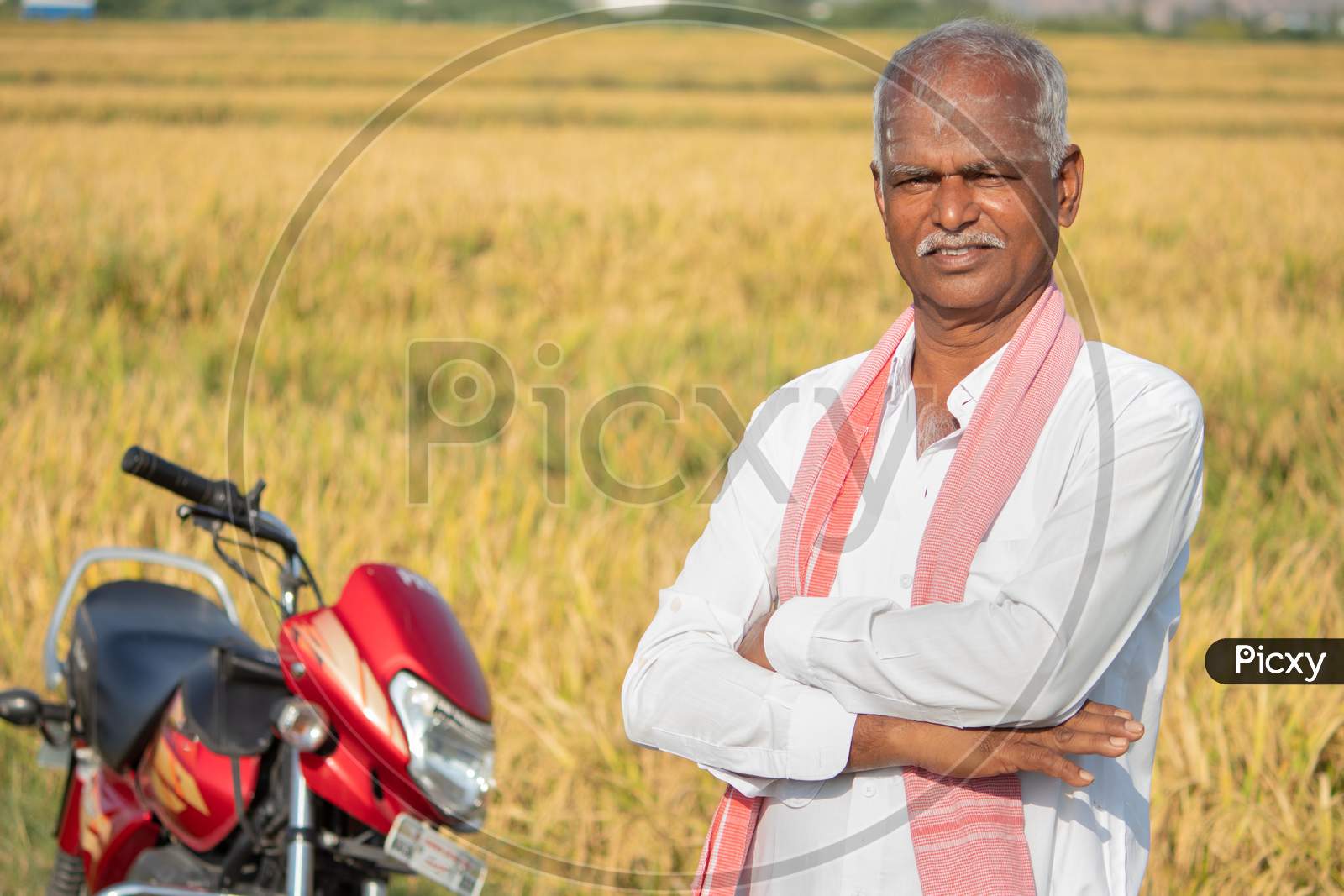 Happy Indian Farmer With Arms Crossed Standing With Bike Infront Of The Agriculture Farmland - Concept Of Good Crop Yield And Farmer Bike Loan.