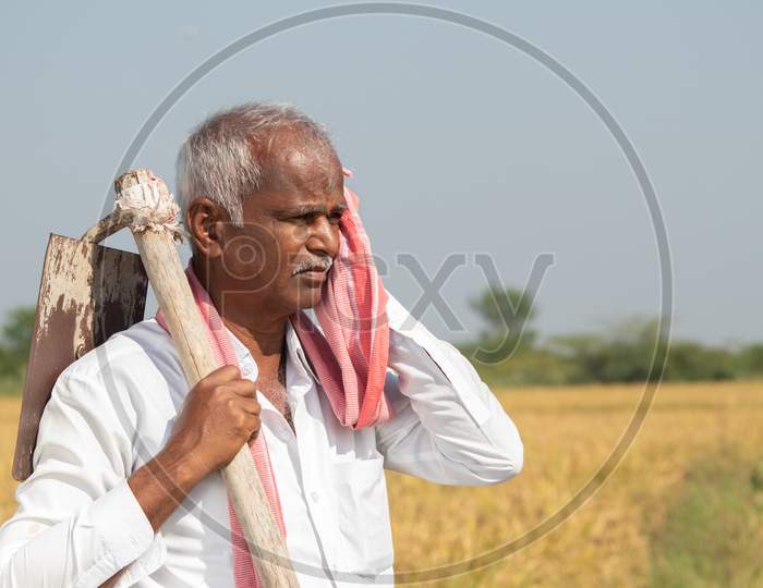 Indian Farmer With Hand Hoe On His Shoulder Holding Towel To His Face Due To High Temperature While Working On Agriculture Land.