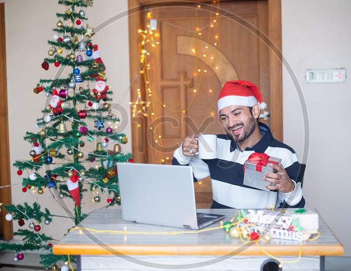 Christmas Celebration 2020, Young Cheerful Indian Man Congratulating Virtually Online On Laptop Computer Showing Gift Present, Pandemic, New Year, Holiday,Winter,December,New Normal.
