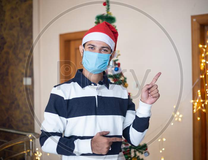 Happy Young Man Wearing Mask And Santa Hat Pointing Finger Posing, Christmas Celebration At Home During Pandemic, Holiday And Winter Season 2020