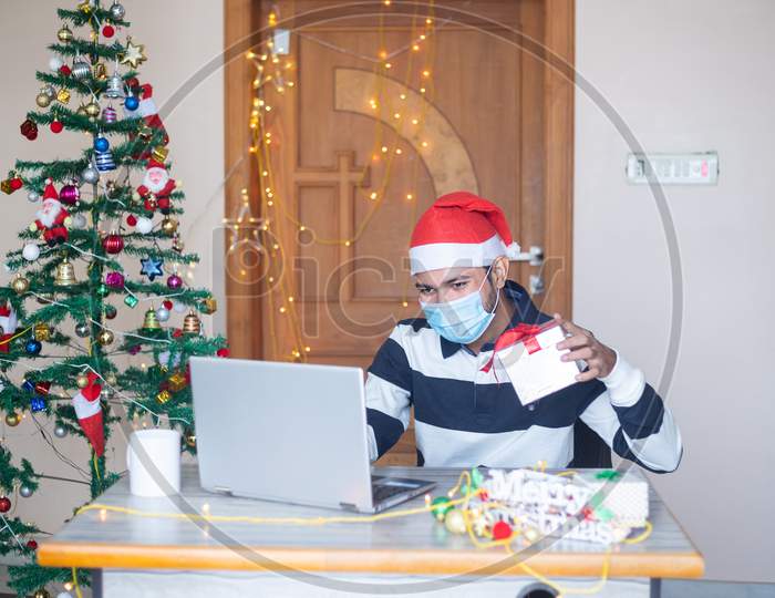 Christmas Celebration, Young Man Wearing Mask Congratulating Virtually Online On Laptop Computer Showing Gift Present, Pandemic, New Year, Holiday,Winter,December 2020,New Normal.