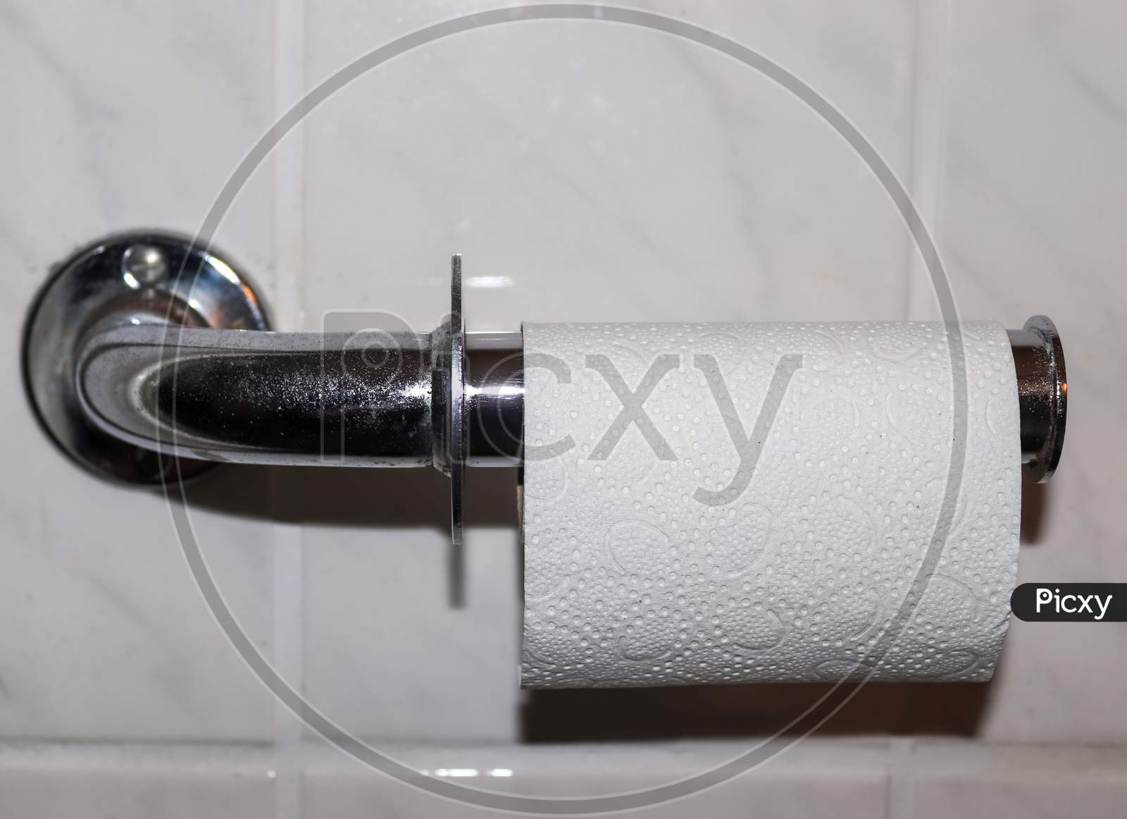 A White Roll Of Soft Toilet Paper Neatly Hanging On A Modern Chrome Holder On A Light Bathroom Wall
