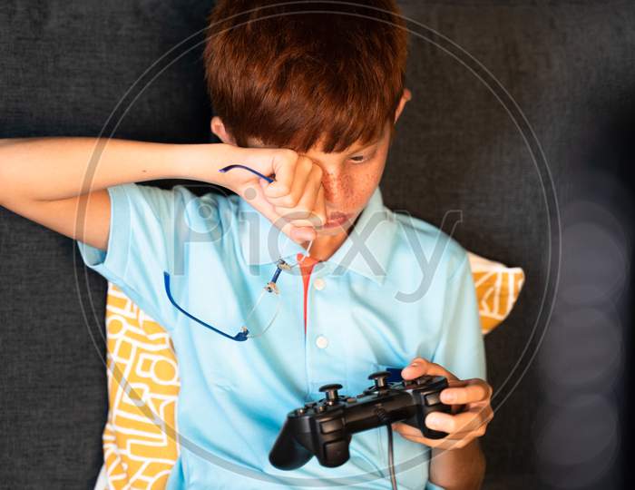 Concept Of Eye Strain Due To Over Night Video Game Play - Teenager Kid Playing Computer Game Using Gamepad During Late Night And Rubbing His Eyes Due To Eye Irritations.