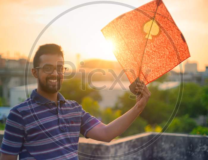 Young Man Holding Aloft Colorful Paper And Wood Kite Against A Blurred Background Setting Sun On The Indian Kite Festival Of Makar Sankranti Or Uttarayana