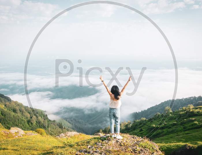 Female Person Out Of Joy Looks To Camera With Spreaded Hands And Scenic Landscape Of Gomismta Mountain Above The Clouds With Sunrise In Backgorun