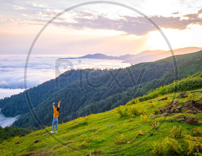 Female Person Out Of Joy Looks To Camera With Spreaded Hands And Scenic Landscape Of Gomismta Mountain Above The Clouds With Sunrise In Backgorun