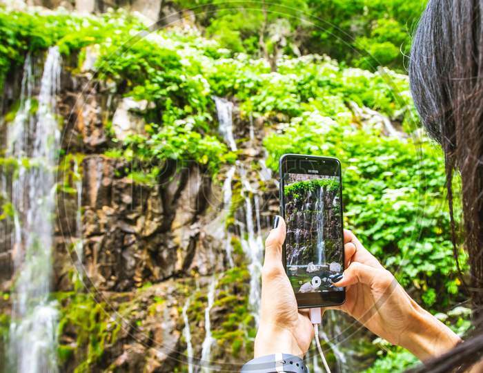 Woman Hand Hold Smartphone And Press Screen To Take A Photo Of Waterfall In Background
