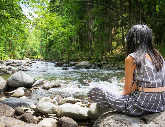 Black Haired Woman Sits On The Rock Looking To River Flow With Green Nature Around