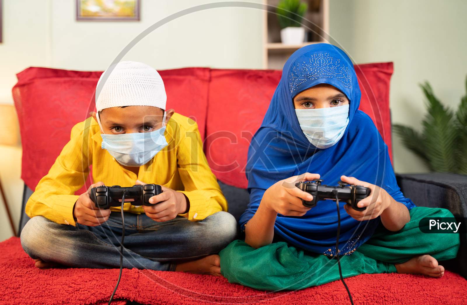Two Muslim Kids With Medical Face Mask Busy Playing Video Game Using Gamepad At Home - Concept Of Kids On Game During Coronavirus Covid-19 Lockdown.