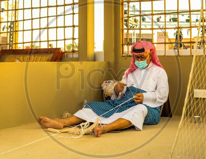 Arabic Fisherman, Repairing Home-Made Fishing Net And Wooden Yacht, Wearing Face Mask And Sits On The Floor Of Traditional Boam