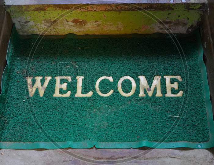 Welcome Home Mat Isolated On A Cement Floor Background. Magnificent Green Colored Logo With Attractive Design.