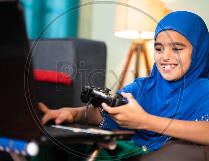 Happy Smiling Muslim Girl Kid Playing Online Videogame On Laptop By Using Gamepad Or Joystick At Home By Wearing Hijab Dress - Concept Of Kid Using Technology And Modern Lifestyle.