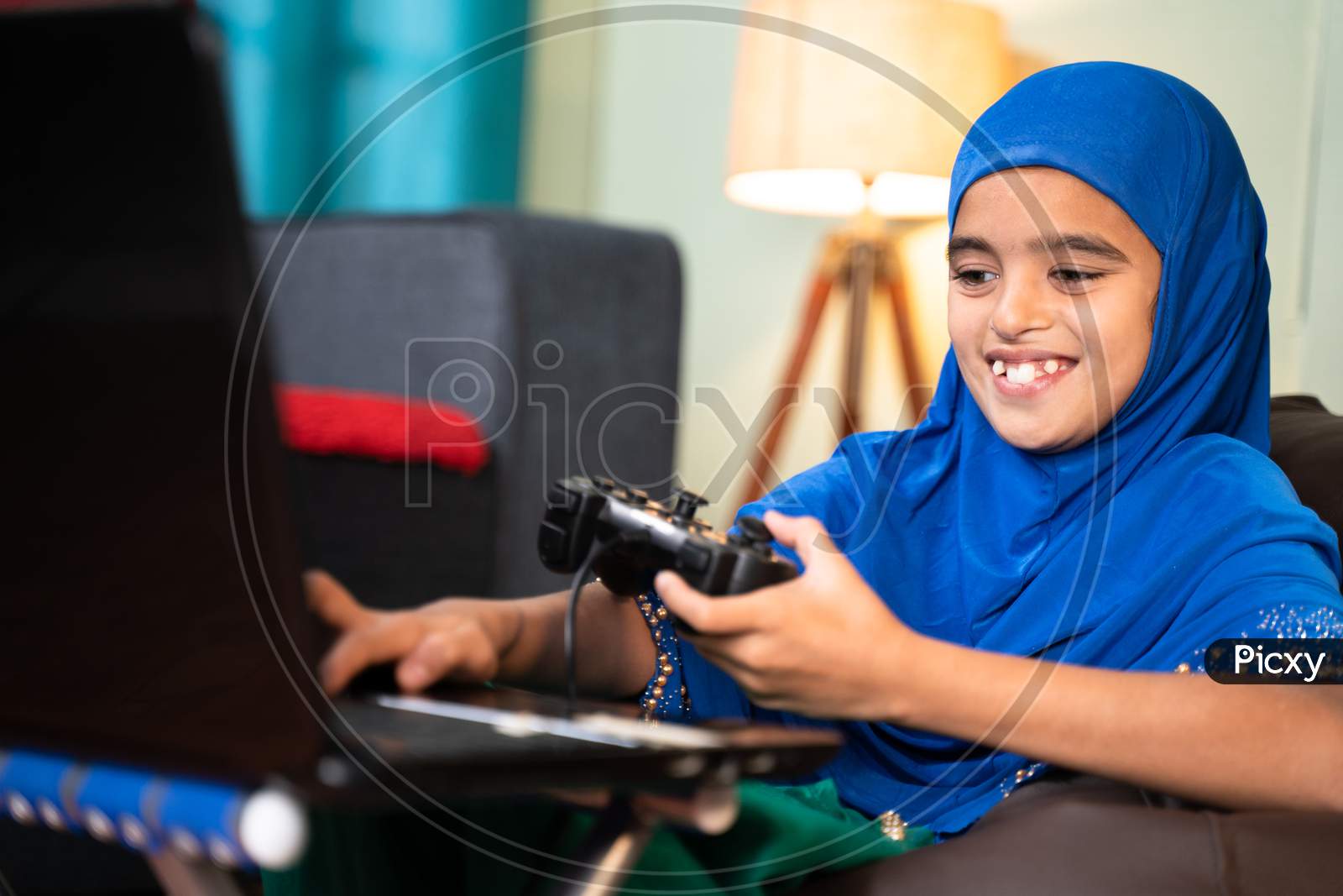 Happy Smiling Muslim Girl Kid Playing Online Videogame On Laptop By Using Gamepad Or Joystick At Home By Wearing Hijab Dress - Concept Of Kid Using Technology And Modern Lifestyle.