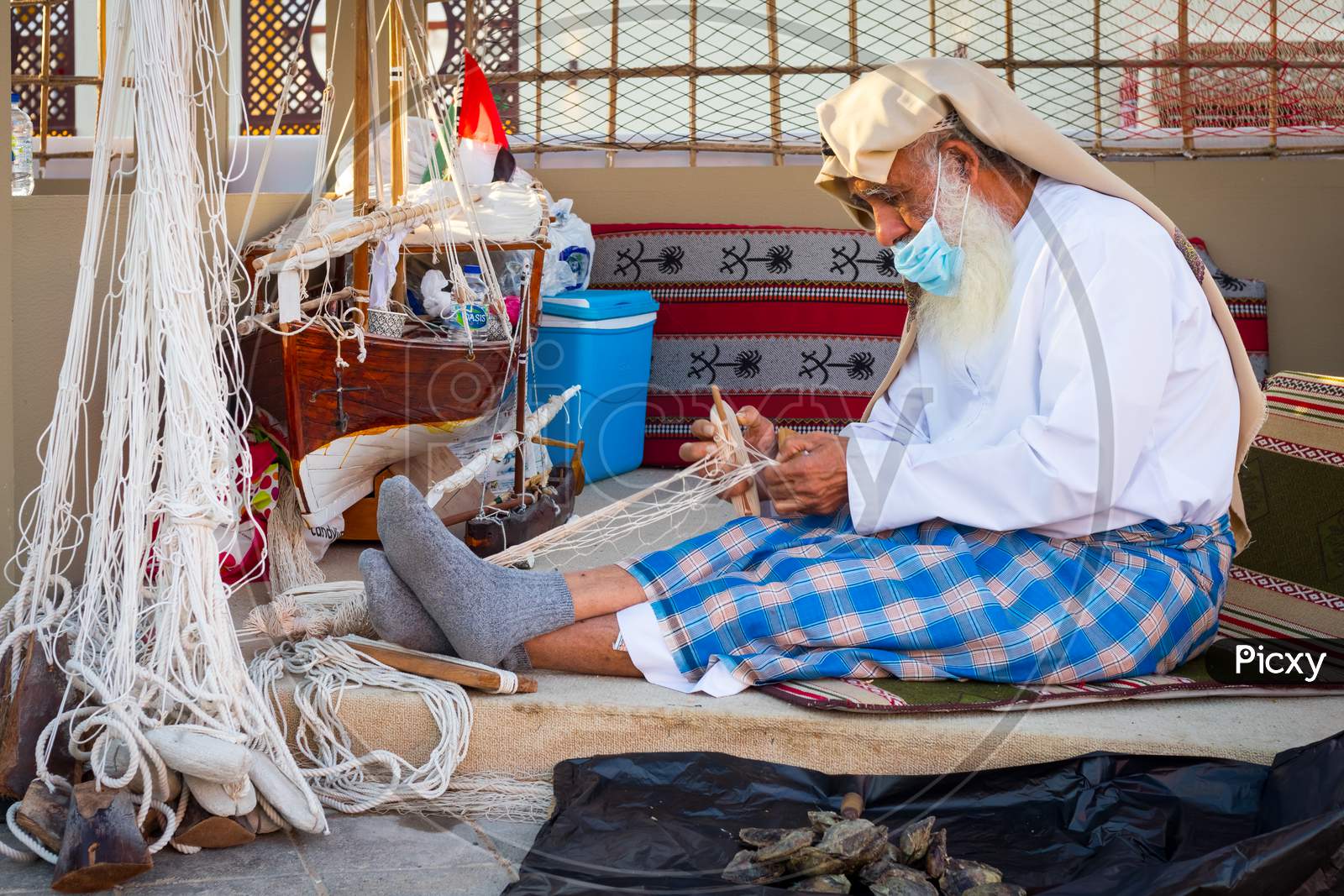 Arabic Fisherman, Repairing Home-Made Fishing Net And Wooden Yacht, Wearing Face Mask And Sits On The Floor Of Traditional Boam