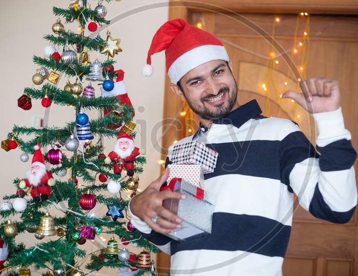 Cheerful Indian Man Wearing Santa Hat Holding Christmas Gift Boxes Or Presents Enjoys Holiday Season At Home, Christmas Tree Background