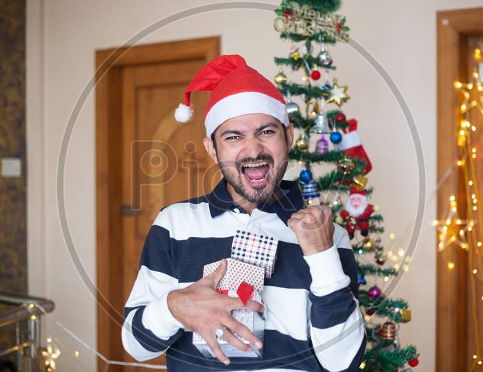 Cheerful Indian Man Wearing Santa Hat Holding Christmas Gift Boxes Or Presents Enjoys Holiday Season At Home, Extreme Happiness,Screams,Crazy Emotions